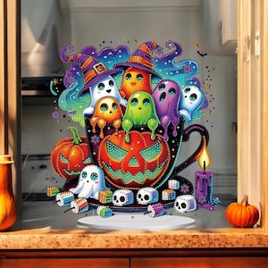 Könnte beinhalten: Eine farbenfrohe Halloween-Dekoration mit einer schwarzen Teetasse, die mit Cartoon-Geistern mit Hexenhüten gefüllt ist. Ein geschnitzter Kürbis und eine brennende Kerze sind ebenfalls vorhanden. Das Design umfasst Totenköpfe und kleine Kürbisse.