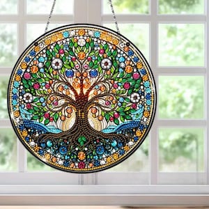 May include: Circular stained glass window art depicting a tree of life. The design features a brown tree with green leaves and colorful gems. The artwork is framed in black and suspended by a chain. The window art is a decorative piece.