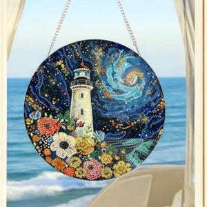 May include: Round, decorative lighthouse art piece with a coastal theme. The artwork features a white lighthouse, colorful flowers, and a starry night sky. The piece is suspended by a silver chain and hangs in front of a blue ocean view.