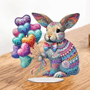 May include: A decorative rabbit figurine covered in colorful, sparkling diamond-like beads. The rabbit wears a patterned bowtie and holds a cluster of heart-shaped balloons in shades of purple, blue, pink, and orange, all featuring a textured, beaded finish.