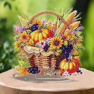 May include: A decorative basket filled with autumn harvest items. The basket contains pumpkins, sunflowers, grapes, and corn. The basket is brown with a clear base, and the background is a blurred green.