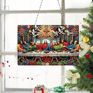 Op de afbeelding: Een rechthoekig glas-in-loodpaneel met het Laatste Avondmaal, met Jezus en zijn discipelen aan een tafel. Het kunstwerk heeft levendige kleuren, waaronder rood, blauw, groen en goud, en is omlijst met een decoratieve rand. Het paneel wordt opgehangen door een zilveren ketting.
