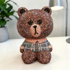 May include: A decorative bear figurine covered in brown, black, and light pink crystals. The bear wears a plaid shirt featuring blue, green, and white tones with a light pink collar and cuffs. The entire surface is textured with small, reflective stones.