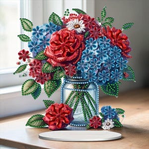 Puede incluir: Un vibrante arreglo floral en un jarrón transparente, con rosas rojas, hortensias azules y margaritas blancas. Las flores están adornadas con detalles brillantes, y el jarrón está sobre una superficie de madera. Un plato de cerámica blanco y redondo está en primer plano.