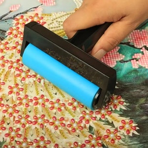 Puede incluir: Primer plano de una herramienta de rodillo azul que se utiliza para presionar los diamantes de imitación en un lienzo de pintura de diamantes. El lienzo presenta un diseño floral con flores rosas y blancas.