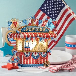 May include: A decorative popcorn stand with red, white, and blue accents, featuring the word "POPOCRN". The stand is decorated with star-shaped and flag-themed elements, and an American flag is in the background. A mug and a plate are also visible.