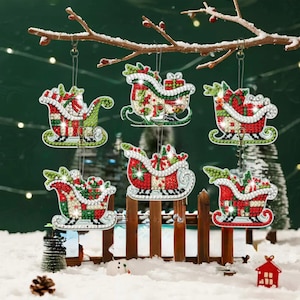 May include: Six hanging Christmas ornaments shaped like Santa sleighs, each decorated with a mosaic of sparkling red, green, and white rhinestones. The sleighs are filled with gift boxes and greenery, suspended by thin clear strings from a tree branch.