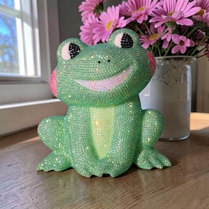 May include: A sparkly green frog figurine with a smiling face, pink cheeks, and black eyes. The frog is covered in glitter and sits on a wooden surface. A vase of pink flowers is in the background.