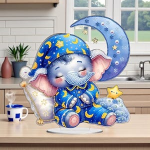 May include: A whimsical acrylic decoration featuring a sleeping baby elephant in blue pajamas and a matching nightcap, resting on a pillow and a crescent moon. The elephant is light blue with pink ears and is surrounded by stars and moons.