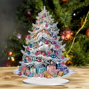 Puede incluir: Un árbol de Navidad decorativo con adornos coloridos y regalos envueltos. El árbol es plateado con detalles en rosa, azul y dorado. Los regalos son de varios colores, incluyendo azul, rosa y dorado. El árbol está sobre un soporte blanco.