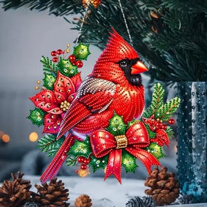 Puede incluir: Un adorno de diamond painting que muestra un pájaro cardenal rojo brillante posado entre hojas de acebo verdes, bayas rojas y un lazo rojo. El diseño está cubierto de pequeñas cuentas facetadas reflectantes y texturizadas.