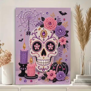 May include: A diamond painting art piece on a light purple background featuring a central skull decorated with white, pink, and purple beads. The design includes two candles, a pumpkin, floral accents, a spider web, and small bats in matching colors.