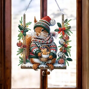 May include: A decorative Christmas window hanging featuring a squirrel wearing a red and green hat, scarf, and sweater. The squirrel holds a basket with a gingerbread man. The hanging is framed with festive greenery, ornaments, and a red bow.
