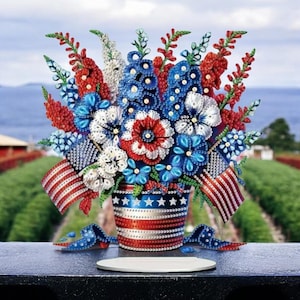 Puede incluir: Un arreglo floral decorativo en rojo, blanco y azul. El ramo está en un cubo con detalles de la bandera estadounidense y dos pequeñas banderas. Las flores están hechas de cuentas y lentejuelas.