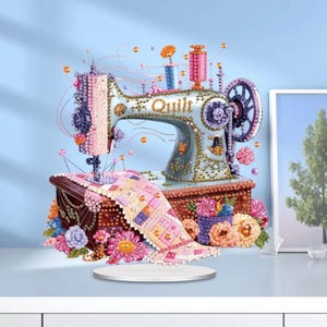 Könnte beinhalten: Eine dekorative Nähmaschine mit dem Wort "Quilt" auf dem Gehäuse. Die Maschine ist hellblau mit goldenen Akzenten und ist von bunten Blumen und Garnrollen umgeben. Auf der Basis liegt ein kleiner Quilt.