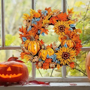 Puede incluir: Una corona otoñal decorativa con calabazas, girasoles y hojas en tonos naranja, amarillo y azul. La corona está adornada con detalles brillantes, ideal para la decoración del hogar en temporada.