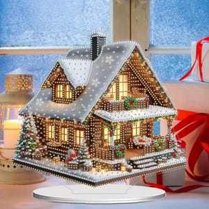 May include: A festive Christmas house ornament with a snow-covered roof and illuminated lights. The house has a chimney, windows, and a small snowman. The ornament is on a white base and is surrounded by festive decorations.