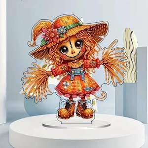Könnte beinhalten: Eine dekorative Vogelscheuchen-Figur mit Diamant-Art-Finish. Die Figur trägt einen orangefarbenen Hut mit breiter Krempe und Blumenakzenten, ein kariertes Kleid mit blauen Trägern und braune Stiefel. Sie hat große dunkle Augen und aufgestickte Details.