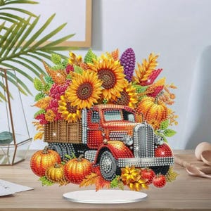 May include: A decorative fall-themed truck figurine. The red truck bed is filled with sunflowers, pumpkins, and autumn foliage. The figurine is embellished with sparkling accents and sits on a clear base.