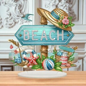Può includere: Un cartello decorativo a tema spiaggia con una freccia blu che recita "BEACH" in lettere scintillanti. Il cartello presenta un cappello di paglia, una stella marina, un pallone da spiaggia e altri oggetti legati alla spiaggia. Il cartello è su una base bianca.