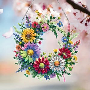 May include: A colorful floral wreath with a variety of flowers in shades of yellow, blue, pink, and red. The wreath is suspended by a silver chain. The background is a soft, blurred image of pink and white flowers.