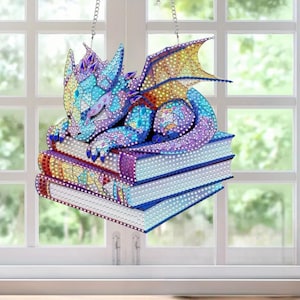 Puede incluir: Adorno decorativo de dragón colorido, descansando sobre una pila de libros. El dragón presenta tonos de azul, morado y amarillo, con un diseño detallado tipo mosaico. Cuelga de una cadena plateada, ideal para ventanas o paredes.