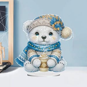 May include: A decorative bear figurine, adorned in a winter hat, scarf, and sweater, holding a golden cup. The bear is primarily white with blue and gold accents. The figurine sits on a clear base, perfect for display.