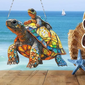 May include: A colorful, decorative turtle ornament with a smaller turtle riding on its back. The ornament is suspended by a chain, with a vibrant mosaic pattern in shades of blue, yellow, orange, and green. The background features a beach scene.
