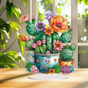 May include: A decorative cactus and flower arrangement in a blue teacup, with a clear stand. The arrangement features various colors of flowers and cacti, including pink, orange, and purple. The teacup is decorated with floral patterns.