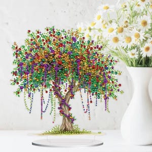 以下が含まれることがあります： 茶色の幹、緑の葉、赤、金、緑、紫のカラフルなビーズアクセントが施された装飾的な木の彫刻。木は透明な台座の上に立っています。背景には白い花瓶とデイジーがあります。