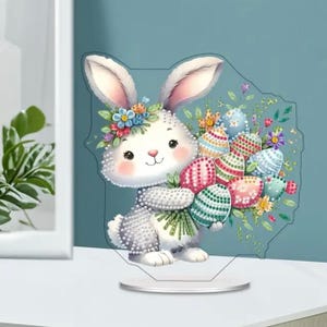 May include: A decorative Easter bunny figurine holding a bouquet of colourful Easter eggs. The white bunny has a floral crown and rosy cheeks. The eggs are decorated with various patterns and colours, set on a clear stand.