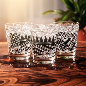 May include: Three clear drinking glasses with black geometric patterns and silver rims, filled with ice. The glasses are arranged on a dark wooden surface, with a plant visible in the background.