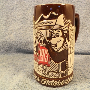 May include: Brown ceramic beer stein with a handle, featuring a cartoon bear in a hat, holding a sign that reads "1973". The stein has a white and brown design with the words "Festes Oktoberfest" around the base.