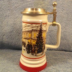 May include: A ceramic beer stein with a metal lid and handle. The stein features a winter scene with a building, a Christmas tree, and figures. The base is red, and the handle and lid are metallic.
