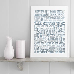 May include: A white framed wall art print featuring a collection of Scottish slang words and phrases in various blue fonts, including the central text TALK SCOTTISH TO ME, displayed alongside a white marble vase and a cylindrical white container.