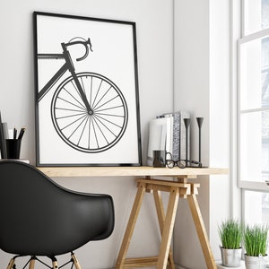 Pu&ograve; includere: Una stampa incorniciata in bianco e nero di una bicicletta con una silhouette nera su sfondo bianco. La stampa &egrave; appesa a una parete bianca sopra una scrivania in legno con una sedia nera, un cavalletto in legno e alcuni libri e piante.