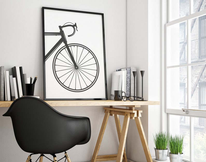 Pu&ograve; includere: Una stampa incorniciata in bianco e nero di una bicicletta con una silhouette nera su sfondo bianco. La stampa &egrave; appesa a una parete bianca sopra una scrivania in legno con una sedia nera, un cavalletto in legno e alcuni libri e piante.