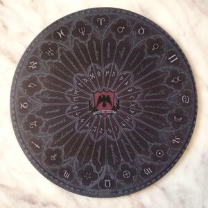 May include: A round, dark gray mat with a gothic stained-glass design. The mat features astrological symbols, runes, and French words. A central emblem displays a crest with a bird. The mat is on a marble surface.
