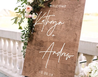 May include: A wooden welcome sign with the names Kathryn and Andrew written in white script. The sign is decorated with greenery and flowers and is displayed on a black easel. The date 10.25.19 is written below the names.