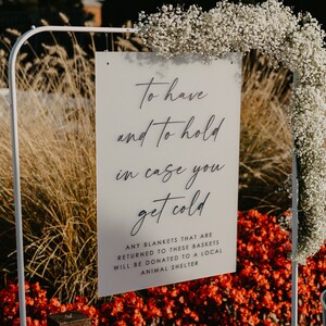 May include: A white sign with black text that reads "To have and to hold in case you get cold. Any blankets that are returned to these baskets will be donated to a local animal shelter." The sign is propped up on a white metal stand. There are two white wire baskets filled with blue blankets.