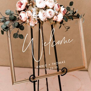 May include: A gold-framed glass sign with the word "Welcome" written in white cursive script. Below the word "Welcome" is the text "Annie + Thomas 10.25.19". The sign is decorated with a floral arrangement of pink and white peonies and eucalyptus.