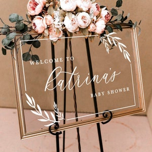 May include: A gold framed clear acrylic sign with white calligraphy that reads "Welcome to Katrina's Baby Shower". The sign is decorated with white floral sprigs and is sitting on a black easel. A bouquet of pink and white flowers with green leaves is sitting on top of the sign.