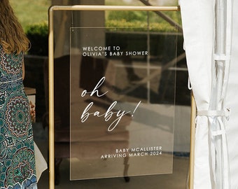 May include: A gold metal stand with a clear acrylic sign that reads "Welcome to Olivia's Baby Shower" and "Oh Baby!" with the text "Baby McAllister Arriving March 2024". The stand is decorated with white candles and green foliage.
