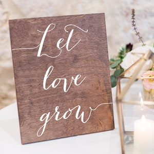 May include: A wooden sign with the text "Let love grow" written in white calligraphy. The sign is sitting on a white surface next to a glass lantern with a lit candle inside.