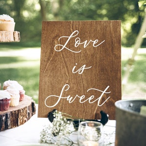 May include: A wooden sign with the text "Love is sweet" in white script on a brown background. The sign is on a stand and there are cupcakes on a wooden tiered stand in the background.
