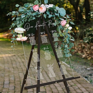 May include: A clear acrylic wedding seating chart with the text "Our Favorite People" at the top. The chart is on a black easel and decorated with greenery and pink flowers.