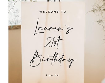 May include: A white welcome sign with black calligraphy text that reads "Welcome to Lauren's 21st Birthday 7.14.24". The sign is on an easel with white flowers and greenery.