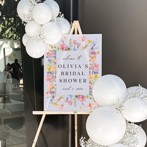 May include: A welcome sign for a bridal shower with a floral design and the text "Welcome to Olivia's Bridal Shower March 5, 2024". The sign is on an easel and surrounded by white balloons with baby's breath.