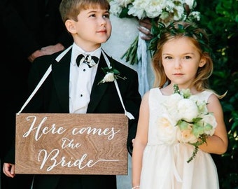 May include: A young boy in a black suit holds a wooden sign that says "Here comes the bride". A young girl in a white dress with a flower crown holds a bouquet of white flowers.