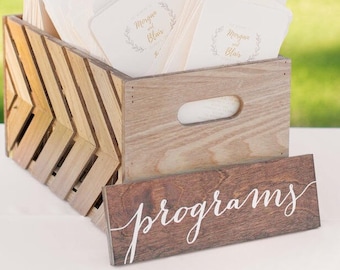 May include: A wooden crate filled with white wedding programs with the names "Morgan & Blair" printed on the front. A wooden sign with the word "programs" in white script is in front of the crate.